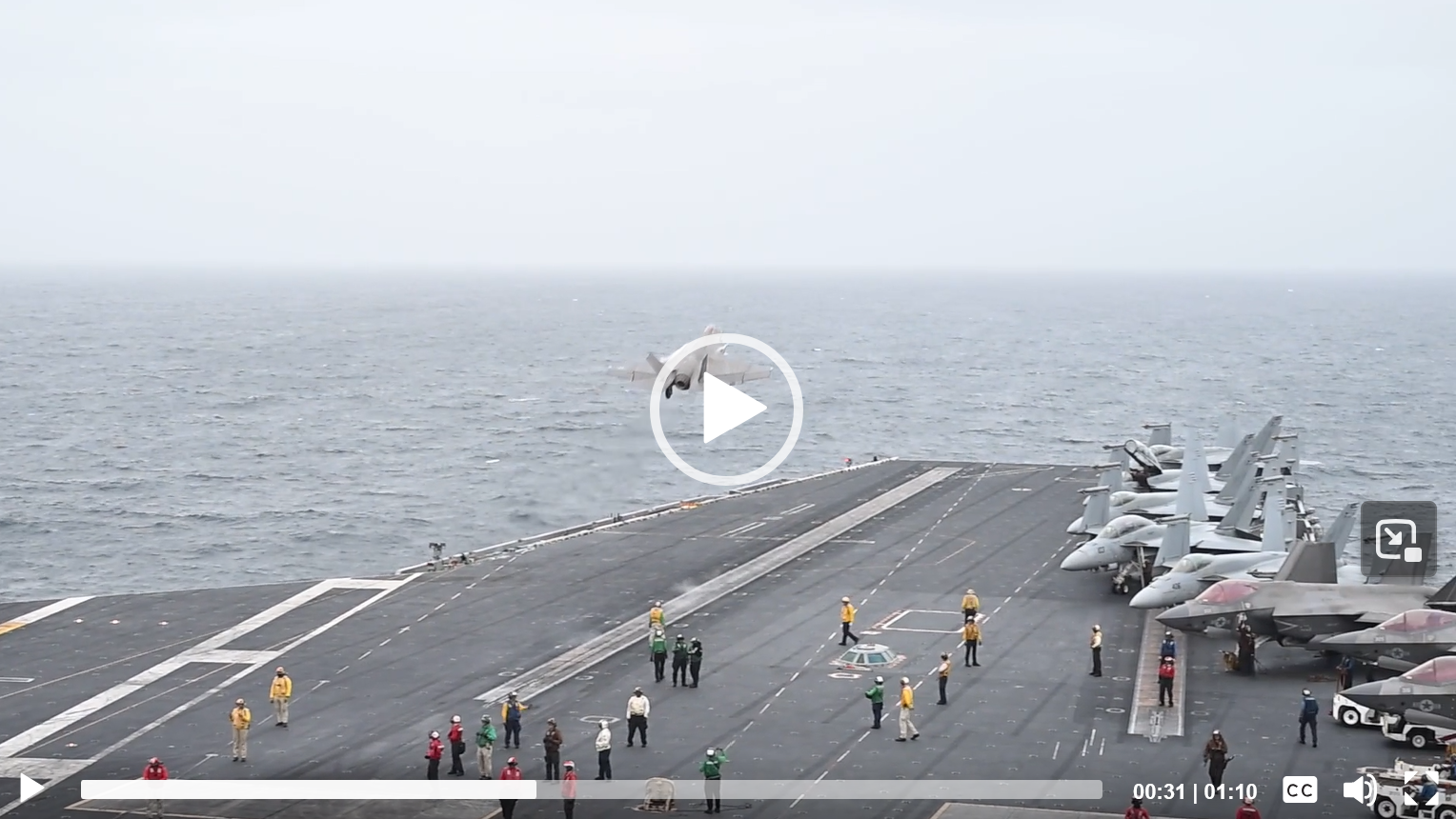 Un avion de combat est en approche d’un porte-avions, avec d’autres avions stationnés sur le pont.